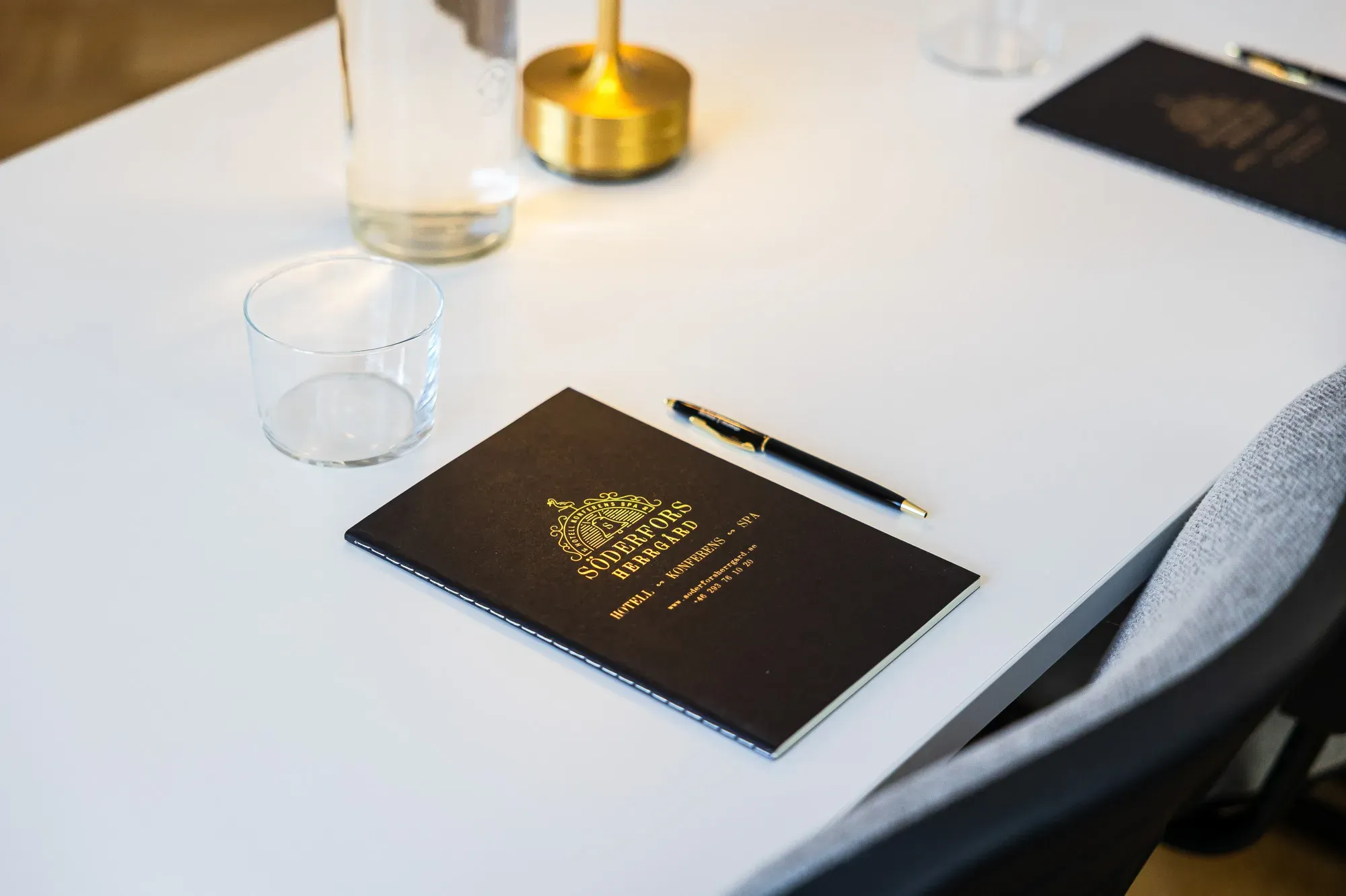Anteckningsbok, penna, vatten och konferenslampa på ett dukat mötesbord.